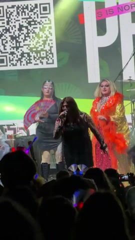 A drag queen performance is taking place on a stage in front of an audience. The stage is decorated with large graphics, including a QR code and stylized fans. Three drag queens are visible on stage. The queen on the left is wearing a black dress and boots and holding a fan. The queen in the center is wearing a black outfit with sheer sleeves and singing into a microphone. The queen on the right is wearing a bright orange sequined outfit with a large fur collar. Later in the video, a queen in a white outfit with a crown and sheer cape takes center stage. The audience is visible in the foreground, with some people holding up phones to record the event. Lighting is dramatic, with stage lights creating colored effects.