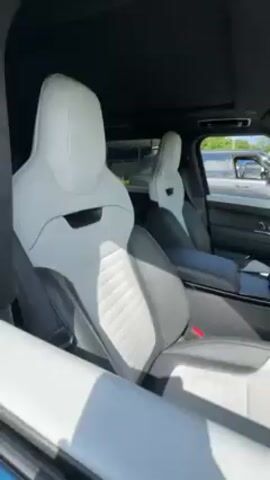 Here is a detailed description of the video:

The video shows the interior of a car, specifically a Range Rover. The focus is on the front seats, which are a two-tone color scheme with white and gray. The seats are designed with a sporty style, featuring prominent side bolsters and unique stitching patterns. The steering wheel is also two-tone, matching the seat colors. The dashboard and other interior elements are black, providing contrast to the lighter colors of the seats and steering wheel. The car has a sunroof, which allows light to stream into the interior. Through the car's windows, one can see other cars parked outside and some greenery, suggesting the car is parked in a lot or near a park.