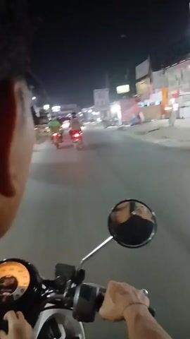 A first-person perspective video shows a person riding a scooter at night on a paved road. The rider's head and shoulder are visible in the foreground. The scooter's handlebars, mirror, and instrument cluster are also visible. The road is lined with buildings and streetlights. Several other vehicles, including cars and motorcycles, are on the road, with their headlights and taillights illuminated. The overall scene is dark, typical of a nighttime urban environment.