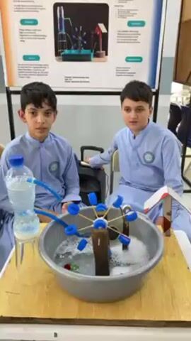 Two young boys, likely students, are seated at a wooden table, presenting or demonstrating a science project. Both boys wear light blue, long-sleeved uniforms or traditional attire (possibly shalwar kameez) with an emblem on the left chest. The boy on the left looks towards the camera with a neutral expression, while the boy on the right looks slightly to his right.

The centerpiece of their presentation is a working model demonstrating a "Water Wheel Free Energy Model," as prominently displayed on a large poster board behind them. The model consists of an inverted clear plastic water bottle elevated on a glass, which feeds water through a blue tube. This tube directs water onto a spinning waterwheel, ingeniously crafted from blue bottle caps attached to thin sticks (possibly toothpicks or skewers) around a central wooden axle. The waterwheel rotates within a gray plastic basin filled with water and foam, indicating it is actively functioning. A small, simple house-like structure with a red roof is placed on the table to the right of the basin.

The poster behind the boys, primarily white with blue and green accents, features diagrams and English text detailing the project's problems, solutions, and methods. The indoor setting, with bright lighting, suggests a school science fair or exhibition. Given the context of Kandahar, Afghanistan, the boys' attire and the project's nature indicate an educational event showcasing student innovation.