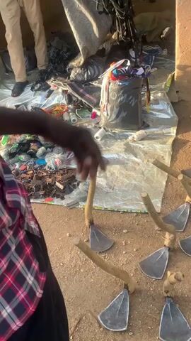 The video opens on a ground-level view of several agricultural tools with metal heads and wooden handles, possibly hoes, arranged in a row on the ground. The camera then pans up and to the left, revealing a makeshift market stall. A person wearing a red and black plaid shirt is partially visible in the foreground, looking towards the stall. The stall itself is covered by a silver tarp, upon which various items are displayed. These items appear to include coils of rope or string in multiple colors, bags of small objects, and other assorted goods. Behind the tarp, a person in a light blue and white striped shirt and tan pants is standing, holding onto a collection of red cords or ropes that are hanging from an unseen structure. The background shows a building wall made of a tan-colored material, and further in the distance, there are glimpses of motorcycles and other people, suggesting an outdoor, possibly rural or semi-urban setting. The overall scene depicts a small market or vendor stall, with a focus on tools and various packaged goods.