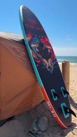 A stand-up paddleboard rests on a sandy beach, leaning against a tan tent. The paddleboard is dark blue with a design of two koi fish in the center and the text "GLIDE WAVE." It has three fins on the bottom and an orange and teal stripe along the edge. In the background, the ocean stretches out under a clear blue sky, with small waves breaking on the shore.