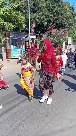 The video shows a procession of people walking down a paved street under a bright, sunny sky. The participants are dressed in colorful, traditional clothing, including some children in elaborate costumes. Some individuals are wearing headscarves. In the background, there are trees, buildings, and a white pole with red and white striped flags. Some people are holding objects, possibly for the procession. The overall atmosphere appears festive.