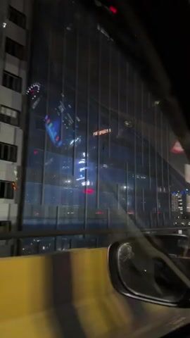 The video shows a night-time view from inside a car driving on a highway. The camera is positioned to capture the windshield and side window. The driver's side mirror is visible. Outside, streetlights illuminate the road and towering buildings in the distance, their windows lit up. Traffic can be seen on the road, with cars and motorcycles appearing as streaks of light due to the motion. A yellow barrier is visible along the side of the road. Inside the car, a "CNG" sticker is affixed to the windshield, along with an "AIRBAG" warning sticker. The car's dashboard and a digital display screen are also visible. The overall impression is of a busy urban environment at night.