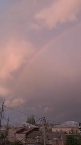 A video displays a pastoral scene of a rainbow arching across a pink and gray sky. Below the sky, a village is visible with rooftops and power lines.