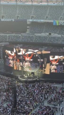 This video shows an aerial view of a large outdoor concert stadium packed with spectators. A band is performing on a circular stage in the center, illuminated by spotlights and large video screens displaying close-ups of the lead singer. The screens show the singer in a circular frame, with other images of him and the band playing instruments. The stadium seating is visible, filled with a diverse crowd of people. Above the stadium, a clear sky with a sunset is visible. The overall atmosphere appears to be energetic and engaging, with the crowd facing the stage and the performers actively engaged in their music.
