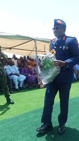 A man in a blue military uniform and sunglasses is walking on a green lawn. He is carrying a large bouquet of greenery wrapped in clear plastic. Behind him, a crowd of people are seated under a white canopy. A person in military fatigues is standing to the left, taking a picture with a phone. The setting appears to be an outdoor event with bright sunlight.