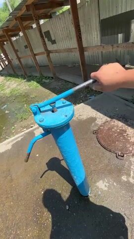 A person's hand is operating a blue water pump. The pump has a long blue pipe and a lever that is being pushed up and down. Water is being pumped out of the pipe and onto the ground. The ground is wet and there is a shadow of a person on the ground. In the background, there is a fence and a building with a wooden roof. The environment appears to be outdoors.