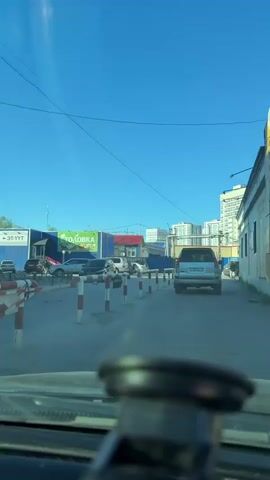 This video captures a street scene in Yakutsk, Russia, under a clear blue sky, suggesting a sunny day. The perspective is from inside a vehicle, looking forward. A silver SUV is the primary moving object in the frame, driving down a road lined with red and white striped bollards. The bollards appear to be part of a traffic control or demarcation system.

In the background, several buildings are visible. To the left, there are low-rise structures with signs that are partially legible. One prominent sign displays the Cyrillic word "СТОЛОВКА," which translates to "canteen" or "cafeteria," and another smaller sign reads "ЭТ-УУТ." Further in the distance, taller, multi-story apartment buildings rise against the sky, indicating a mix of commercial and residential areas.

The road surface appears to be asphalt or a similar material. A few other vehicles are parked or moving in the distance, and a person can be seen walking on the right side of the road. The overall impression is of an ordinary day in an urban environment, with the clear weather contributing to good visibility. The presence of the parked cars and the general layout suggest a commercial district or a street leading into one.