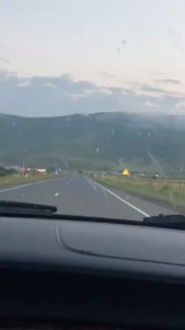 The video appears to be taken from inside a car driving on a highway. The sky is overcast with a hint of yellow in the distance, suggesting either sunrise or sunset. Mountains are visible in the background, partially obscured by clouds or mist. The road ahead is clear, with a few other vehicles visible in the distance. A side mirror of the car is also visible in the foreground, reflecting the sky and the passing landscape. The overall impression is a scenic drive through a rural or mountainous area during twilight.
