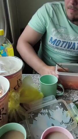 This media file appears to capture a meal on a train, interspersed with views of the passing scenery. The primary subject is a man, wearing a light green t-shirt with the text "Competition MOTIVATION THE FIRST STEP TO BEAT YOUR LAZINESS" printed on it. He is seated at a table laden with food containers, a green mug, and a bottle of Lipton iced tea. The meal appears to consist of some sort of stew or soup, as well as what looks like pastries or bread.

The scene transitions to showcase the view from the train window, revealing a lush, green landscape with abundant trees and vegetation. The sky is visible, dotted with clouds, suggesting it is daytime. There are some power lines and a utility pole visible in the background of the train view. The overall impression is one of travel and a simple meal enjoyed en route. The man's expression is neutral, suggesting a relaxed or perhaps contemplative mood during his journey. The context provided indicates the location is Syrt, Russia.