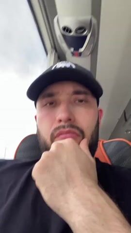 A man with a beard and mustache, wearing a black baseball cap and a black t-shirt, is seated inside what appears to be a bus or passenger vehicle. He looks directly at the camera with a neutral, possibly contemplative or bored, expression. His right hand rests on his chin, occasionally adjusting its position. Behind him is an orange and grey patterned seat, and to his left, a window shows a cloudy sky. Above him, an overhead panel with vents is visible. The internal structure of the vehicle and the man's posture suggest he is a passenger. No clear text is legible on his clothing.