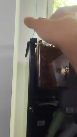 A person's hands are visible as they remove a clear plastic container filled with coffee beans from a black machine. The container is tilted, and some of the beans are spilling out as it is being removed. The machine appears to be some kind of dispenser or coffee maker. In the background, a blurry image of a person's face is visible, as well as glimpses of a window and some greenery.