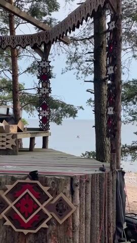 A wooden structure with traditional folk art decorations is in the foreground. In the background, the calm sea is visible under a cloudy sky. Two people are on a paddleboard in the distance.