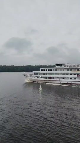 A white cruise ship with red trim sails across dark, choppy water under a cloudy sky. The ship has multiple decks with numerous windows. The shoreline is visible in the distance, lined with trees. The camera angle is from a slightly elevated position, looking towards the ship.
