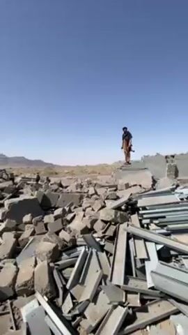 The video shows a person standing on a pile of rubble in a desolate landscape. The person is wearing a long, dark garment and is facing away from the camera. The rubble consists of broken concrete blocks and pieces of metal framing. In the background, there is a barren landscape with distant mountains under a clear blue sky. The video is shot from a low angle, emphasizing the scale of the destruction.
