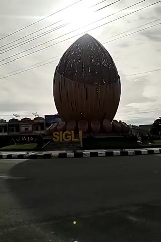 The video shows a large, brown, oval-shaped monument in the middle of a traffic roundabout. The monument resembles a coconut, with a latticework pattern on top. The word "SIGLI" is spelled out in large yellow letters at the base of the monument. The roundabout is surrounded by a black and white checkered curb. The sky is overcast. There are buildings and trees in the background. The video is stationary.