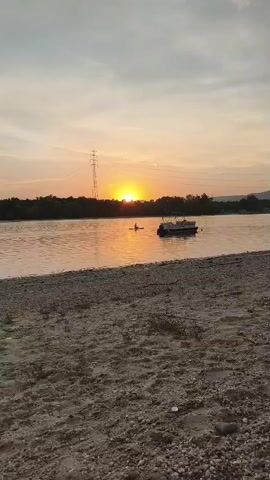 The video shows a sunset over a body of water. In the foreground, there is a sandy and pebbly beach. The water is calm and reflects the orange and yellow hues of the sunset. In the middle ground, on the water, there is a person in a kayak and a small boat. In the background, there is a tree line and hills. A tall electrical tower is visible in the distance on the left side of the frame. The sky is a gradient of orange, yellow, and gray, with some clouds.