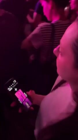 This media file captures a live concert performance at night. The scene is dominated by vibrant pink and purple stage lighting, creating a dramatic and energetic atmosphere. In the foreground, the audience is visible, with some individuals holding up cell phones, suggesting they are recording or photographing the event. One person in the foreground is holding a pink fan.

The focal point of the video is the stage, where a female performer with short, reddish hair is singing into a microphone. She is wearing a dark sleeveless top, shimmering sequined shorts, black tights, and platform boots with bright orange heels. Her energetic poses and gestures indicate she is actively engaging with the audience. Another figure, likely a band member, can be seen on the stage in the background.

The stage is equipped with professional lighting rigs and sound equipment, typical of a concert venue. Smoke or fog machines are being used, adding to the theatrical effect of the lighting.

The video appears to be taken in Kragujevac, Serbia, as indicated by the context provided. The time of day is clearly night, due to the artificial stage lighting and the darkness of the surroundings. The overall mood is exciting and celebratory, reflecting the energy of a live music performance. No visible text is discernible within the frames.