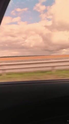 The video shows a view from inside a car looking out the window at a highway. The sky is filled with pink and white clouds. In the distance, there is a field of orange-brown color, possibly farmland. A red car passes by on the highway. The car window frame is visible in the foreground.