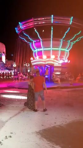 The video shows a fairground at night. The primary focus is a large swing ride decorated with blue and white lights. In the foreground, a couple stands looking at the ride. The woman is wearing a long, dark floral dress, and the man is in a red t-shirt and blue shorts. The ground is sandy and uneven, with some fallen leaves. Further back, other amusement rides and attractions are visible, including a carousel with colorful lights and a ride with a dancer figure. The overall atmosphere is festive and energetic, illuminated by the bright lights of the rides.
