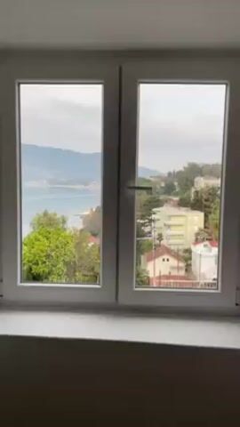 The video shows a view from inside a room through a closed double-paned window. The window is white framed. A hand reaches forward and opens the right side of the window. The view through the window reveals a coastal town with buildings, trees, and a body of water in the background, framed by mountains. The sky is overcast.