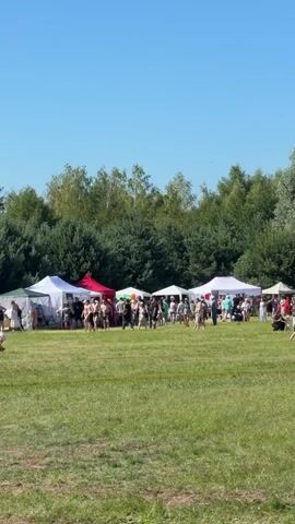 The video shows an outdoor gathering on a sunny day. There are several white and red tents set up on a grassy field. Many people are walking around and socializing. Some people are sitting in lawn chairs. There is a large group of trees in the background. In the foreground, a few people are visible walking towards the camera. One person is wearing a black dress. There are colorful ribbons attached to poles in the middle of the field.