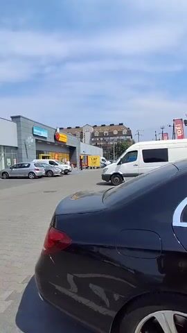 This video showcases a commercial strip mall on a sunny day in Ząbki, Poland. The scene is bustling with activity, featuring several cars parked in the lot and people walking in and out of the stores. The sky is blue with scattered white clouds, indicating pleasant weather.

The video pans across the storefronts, revealing the names of various businesses. Prominently displayed are "Rossmann," "pepco," and "empik." A white van is parked in the foreground, and a black sedan is partially visible in the lower right corner. The ground is paved with gray interlocking bricks, typical of outdoor shopping areas.

The time of day appears to be late morning or early afternoon, given the bright sunlight and the shadows cast by the buildings and vehicles. The overall atmosphere seems to be one of everyday commerce and activity, with no specific event or emotion being highlighted. The setting is clearly a functional shopping center, catering to the local community. The visible text on the signs provides clear identification of the retail establishments.
