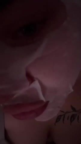 A woman wearing a white sheet mask and a gray tank top is shown in close-up. She has a leaf tattoo on her upper chest. The lighting is dim, creating shadows and highlighting her face. The woman appears to be indoors, possibly in a bedroom, as indicated by the soft lighting and what looks like bedding in the background. She shifts her position slightly throughout the short video, looking towards the camera at times.