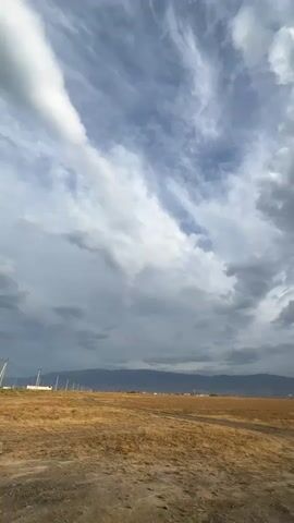 A still, vertical video captures a vast, open landscape under a dramatic sky. The foreground is a dry, golden field with a dirt path winding through it. In the distance, a range of mountains stretches across the horizon, their peaks softened by haze. The sky is a dynamic mix of clouds, with wispy cirrus clouds high above, streaked with white against a blue background, and heavier, darker cumulus clouds lower down. Sunlight breaks through in some areas, casting subtle rays. The scene is devoid of people and any discernible activity, emphasizing the natural environment.