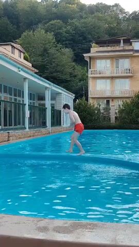 A young person in red shorts stands on the edge of a swimming pool, facing away from the camera and towards the water. Behind them, a building with a light-colored facade and balconies is visible, surrounded by green trees. The pool itself is filled with bright blue water, with some ripples and white foam indicating movement. Another person is partially submerged in the pool to the right of the frame.
