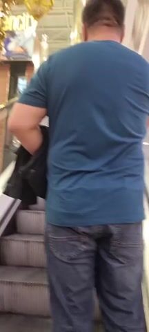 The video shows a view from an escalator inside a shopping mall. A person's back is visible on the escalator, facing away from the camera. Below the escalator, an ice rink is set up with an ice skating area, surrounded by barriers. Several orange cones are placed on the ice. In the background, shops are visible on different levels of the mall, with escalators and people moving around. The lighting suggests the mall is open and active.