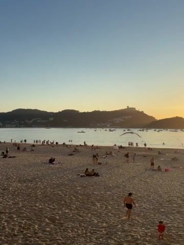 A wide shot captures a crowded beach at sunset. The sand is a warm golden color, with many people scattered across it, some lying down, others walking or standing in groups. The ocean stretches out beyond the beach, dotted with a few boats. In the background, a hilly landscape is visible, with buildings perched on top of one of the hills. The sky is a clear, soft blue, with the sun setting on the right side of the frame, casting a warm glow over the scene. The overall atmosphere appears relaxed and enjoyable, with people taking advantage of the pleasant evening weather.