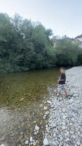 A young boy in a black t-shirt and patterned shorts stands on a rocky riverbank, looking towards the water. He is holding something in his right hand and appears to be in motion, possibly about to throw something. The river is shallow and clear, with visible pebbles on the riverbed. Lush green trees line the opposite bank, and a few buildings are visible in the distance. The sky is overcast.