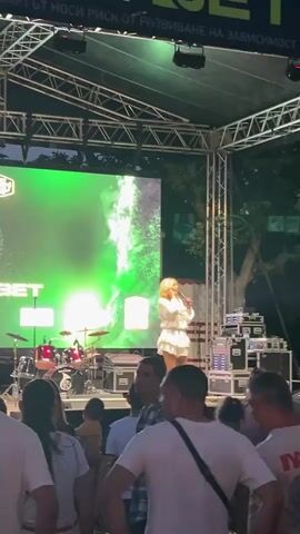 This is a vertical, medium shot video taken from behind an audience watching a performance on an outdoor stage at night. A blonde female singer in a white dress is standing center stage, holding a microphone and singing into it. She is facing the audience. Behind her, a large screen displays green abstract visuals and text. To the left of the screen, a drum set is visible. On the right side of the stage, audio equipment is stacked. The audience members in the foreground are mostly wearing white t-shirts. The stage is set up under a metal truss structure with lighting fixtures. Trees are visible in the background.