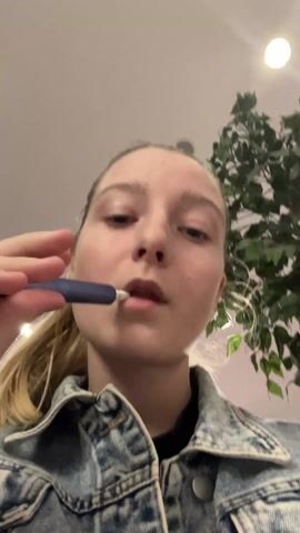 A young, fair-skinned woman with blonde hair pulled back in a ponytail is the focal point of the video. She is wearing a blue denim jacket and a black top underneath. The camera is positioned at a low angle, looking up at her face. She is shown taking a puff from a vaping device, then exhaling a cloud of white smoke. She smiles. In the background, there is a ceiling with recessed lighting and a faux green leafy plant. The overall scene appears to be indoors.