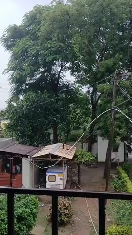 The video shows a view from a balcony overlooking an outdoor area, possibly a courtyard or street, during what appears to be a rainy day. In the foreground, a black railing is visible. Beyond the railing, there are manicured bushes and trees. In the middle ground, there is a small shed with a bamboo-like roof covering a white generator. A utility pole with numerous electrical wires stretches across the scene, connecting to structures in the background. A paved path leads to a metal gate, which is open. Beyond the gate, there are buildings and more trees. The ground appears wet, indicating recent or ongoing rainfall. The overall atmosphere is subdued due to the overcast sky and rain. There are no people clearly visible in action, though a few figures might be present near the gate in the distance.