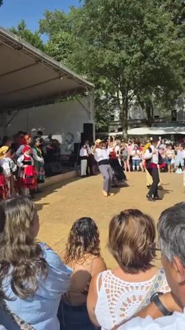 The video shows an outdoor festival with a stage set up. A group of people in traditional folk costumes are dancing. One couple in the foreground is dancing in sync, with the man wearing a straw hat and the woman in a long, colorful skirt. The background shows more dancers, musicians on stage, and a crowd of spectators. The environment appears to be a sunny day in a town square, with trees and buildings visible in the background.
