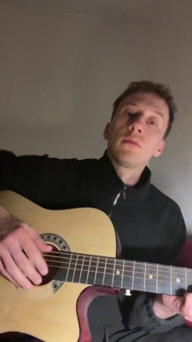 A man plays an acoustic guitar while sitting on a red sofa. He wears a black zip-up sweater. He looks directly at the camera and raises his middle finger. The wall behind him is plain.