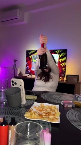 A woman excitedly pops the cork off a champagne bottle in an indoor setting. She is sitting at a table with several items on it, including a smartphone, a mug, glasses, a plate of potato chips, cigarettes, and vaping devices. In the background, a television displays the words "THINK ABOUT IT". The room has an air conditioner, and the walls are illuminated with purple and blue lights. The woman is visibly happy, with a wide smile and arms raised in celebration.