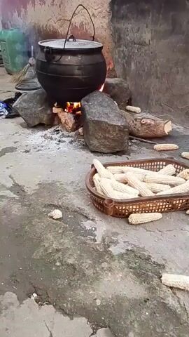 A black pot is suspended over a fire by large rocks. A pile of wood is burning under the pot. In front of the fire, a brown basket overflows with husks of corn. Several loose husks are scattered on the ground around the basket. In the background, a rustic building with a rough wall is visible.