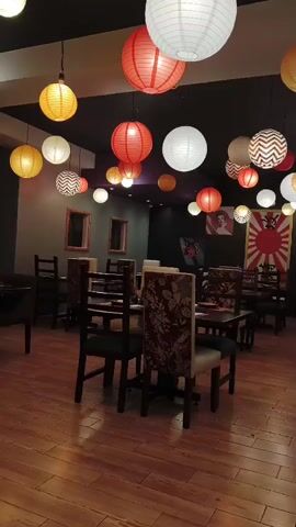 The video shows the interior of a restaurant. The floor is made of light brown wooden planks. Numerous colorful paper lanterns hang from the ceiling, including white, red, and yellow lanterns. There are several tables and chairs in the room, arranged for dining. Some chairs have floral patterns, while others are plain. The walls are painted in dark gray and black, with framed mirrors on one wall. At the back of the room, there's a mural featuring a Japanese flag. The overall atmosphere is casual and inviting, with a touch of Asian-inspired decor.