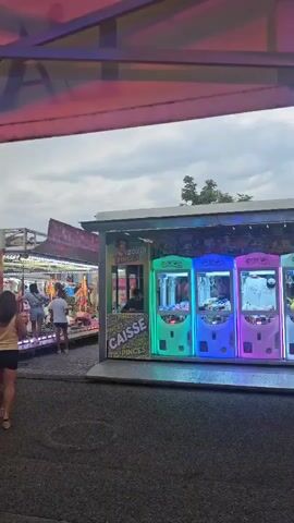 A group of people are walking through a midway at what appears to be a fair or amusement park. In the foreground, a woman in a yellow top and black skirt walks away from the camera. To her left, a red attraction with the word "FURIOUS" in large yellow letters is visible. To her right, a row of brightly lit arcade claw machines, each in a different color, are set up. Several people are standing near the machines and other attractions in the background. The sky is cloudy and the lighting suggests it is late afternoon or early evening.
