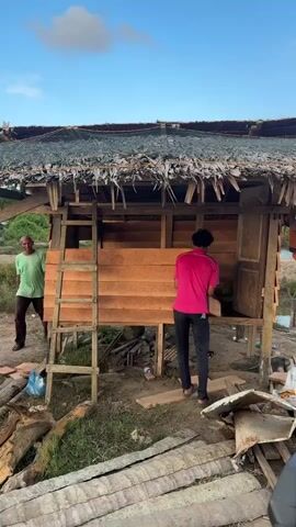 Two men are seen working on a wooden structure. One man, wearing a green shirt, stands to the left of the structure, looking towards the other man. The second man, wearing a bright pink shirt, is positioned in front of the structure, appearing to be working on the wooden planks. The structure itself is elevated on stilts and has a thatched roof. There are piles of wood and debris surrounding the structure, suggesting construction or renovation work is in progress. The environment appears to be outdoors with a clear sky and vegetation visible in the background.