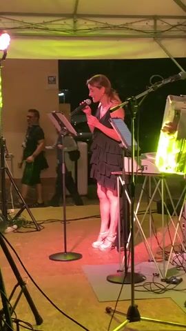 A female singer in a black ruffled dress and white high heels stands on a stage under a white tent, holding a microphone and singing. She has a music stand with a tablet in front of her. To her right, an accordion player is visible, along with a keyboard and other musical equipment. In the background, two people are walking, and a white cabinet is behind the singer. The stage is lit with pink and purple lights.