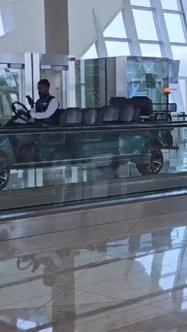 A man wearing a dark vest over a white shirt and dark pants drives a dark green golf cart with multiple black seats on an escalator. He is holding a phone in his left hand and appears to be looking at it. The golf cart has tinted windows and is moving forward. Other passengers are visible in the background. The environment appears to be an indoor public space with large windows and modern architecture. The floor is reflective.