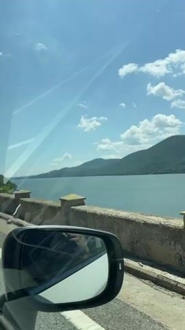 The camera is positioned inside a moving vehicle, looking out the passenger window. The side mirror of the vehicle is visible in the bottom left corner. The vehicle is traveling along a road that runs parallel to a large body of water, likely a river or lake, with green, tree-covered mountains in the distance on the opposite side. A stone wall or barrier separates the road from the water. The sky is bright blue with scattered white clouds. The sun is shining, creating reflections and glare on the car window. The view changes slightly as the vehicle moves forward, showing different sections of the water, mountains, and the roadside barrier.