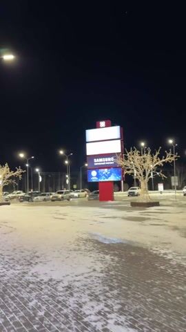 The video depicts an outdoor scene at night, possibly a parking lot or open space in front of a commercial building. The ground is covered in a mix of snow and pavement. Several cars are parked in the lot, illuminated by streetlights. A large, red sign structure is visible, featuring several illuminated panels. One panel displays the Samsung logo. Flanking the sign structure are two trees decorated with strings of lights, giving them a glowing appearance. Additional streetlights are spaced throughout the area. A sign advertising auto services is also visible on the left. The sky is dark and clear.