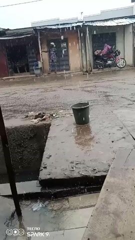 The scene is a rainy day on a street with small shops on either side. The ground is wet and there is standing water on the road.  A dark green bucket sits on a concrete surface next to a drainage area. A man wearing a dark shirt, shorts, and sandals walks across the street while wearing headphones. A person in a military-style uniform stands under the awning of one of the shops. A red motorcycle is parked in front of another shop. The video is recorded with a phone; the phone brand and model is watermarked in the corner.