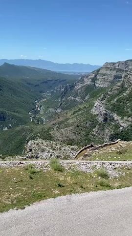 The video captures a panoramic view of a lush green valley with a winding river. The valley is carved between steep, rocky mountains, some of which are covered in sparse vegetation. The sky is clear and blue, indicating fair weather. In the foreground, there's a gravel path and a stone wall, suggesting a viewpoint or a hiking trail. On top of the wall, a group of people can be seen observing the landscape. The river flowing through the valley appears to be a vibrant turquoise. In the distance, the valley opens up to a larger body of water, possibly a lake or the sea, surrounded by more mountains. The overall scene is one of natural beauty and tranquility, showcasing a mountainous landscape with a river and distant water body.